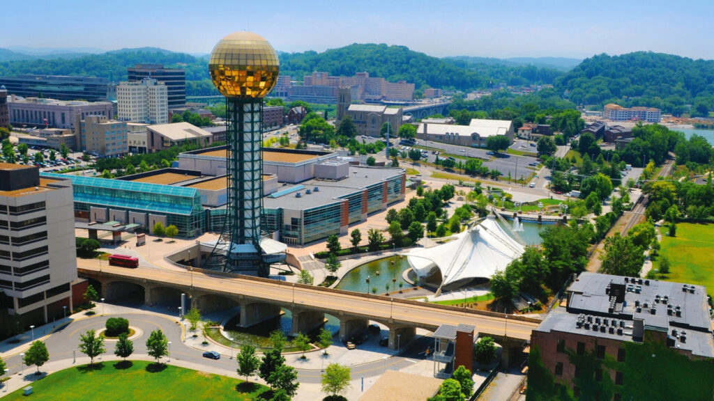 Aerial view of downtown Knoxville, Tennessee, featuring the iconic Sunsphere tower overlooking the city, with surrounding buildings, bridges, and green spaces along the waterfront.