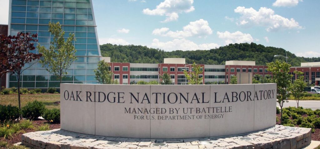 Entrance sign for Oak Ridge National Laboratory with modern campus buildings and landscaped grounds in the background.