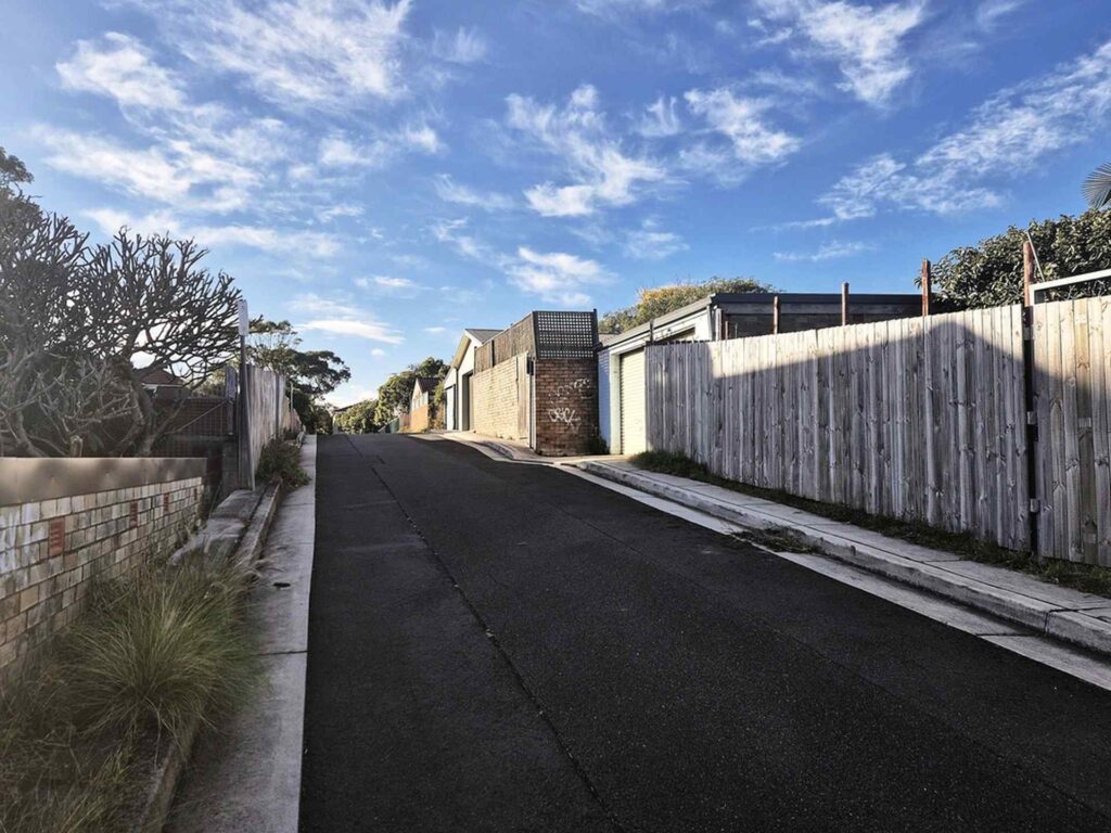 Freshly paved private laneway with smooth asphalt surface, bordered by residential fences, garages, and sidewalks under a partly cloudy sky.