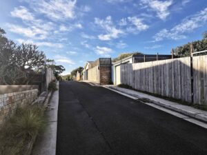 Freshly paved private laneway with smooth asphalt surface, bordered by residential fences, garages, and sidewalks under a partly cloudy sky.