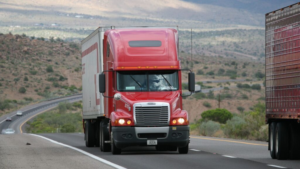 Red semi-truck driving on an asphalt access road illustrating heavy winter traffic wear.